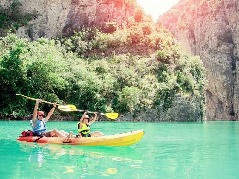 Man en kind in een kano, juichend met hun peddels in de lucht, omringd door turquoise water en rotsen.